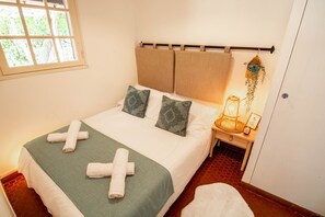 A neatly made bed with a green blanket and two pillows, a wooden headboard, a bedside table with a lamp, and a window with a view of trees.