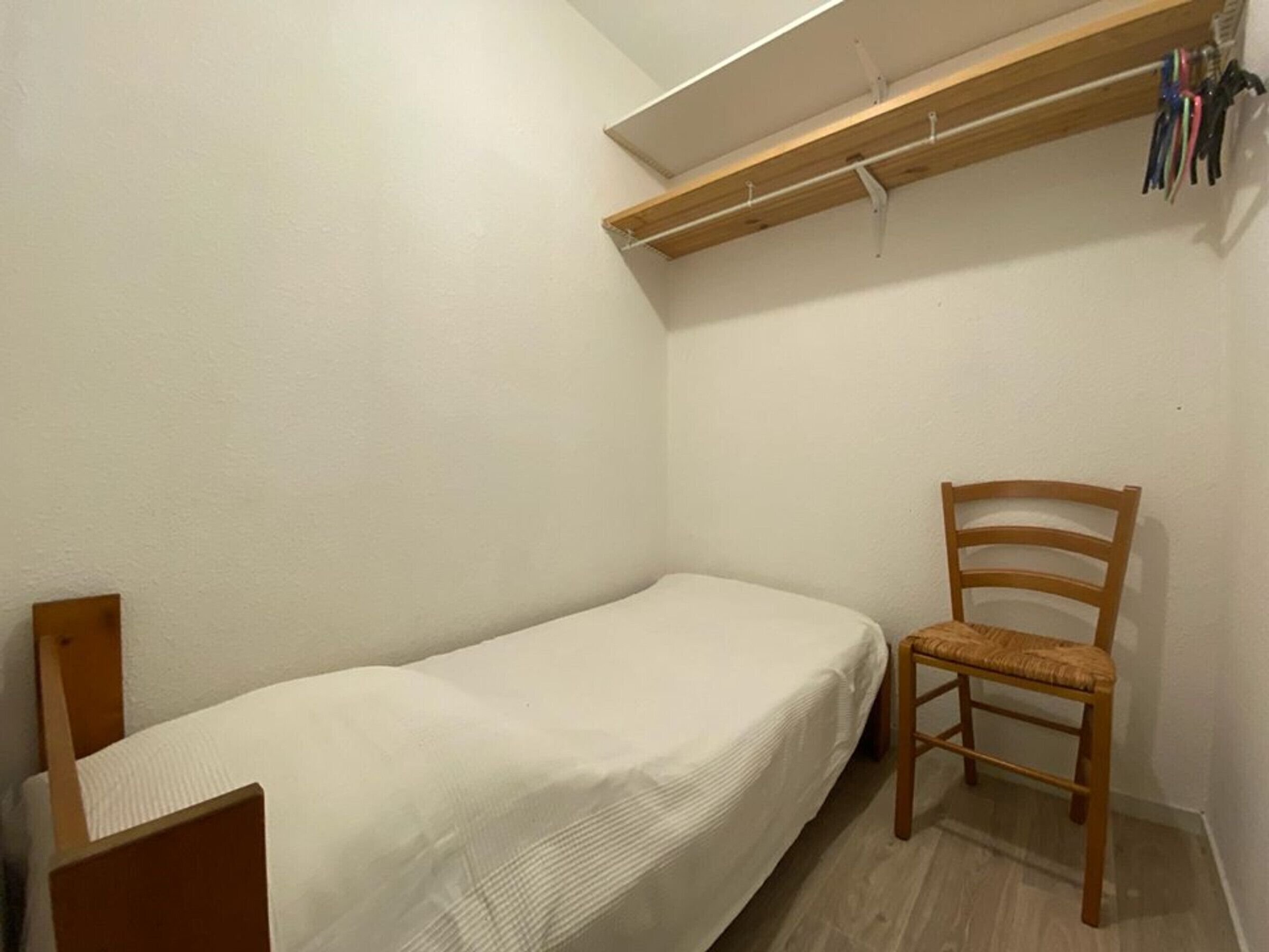 Une petite chambre avec un lit simple, une chaise en bois et une étagère au-dessus du lit.