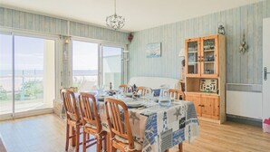 A dining area with a table set for four, wooden chairs, a chandelier, a view through large windows, and a wooden cabinet with glass doors. - 3-bedroom apartment with large balcony and garage near the casino (Saint-Jean-de-Monts)