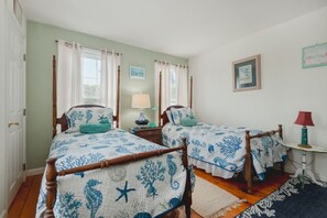 Una habitación con dos camas, cada una con estructura de madera y ropa de cama estampada en azul. Hay dos mesitas de noche con lámparas, una ventana con cortinas y una alfombra en el suelo.