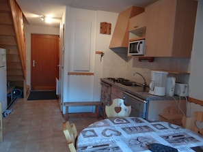 Cozinha compacta com fogão, pia e micro-ondas. Há um banco de madeira e uma mesa com toalhas de coração. Uma porta de madeira e escada são visíveis ao fundo.
