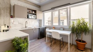 Cozinha moderna com micro-ondas embutido, pia e área de jantar com mesa e cadeiras brancas. Há uma planta em vaso e uma janela com vista para edifícios.