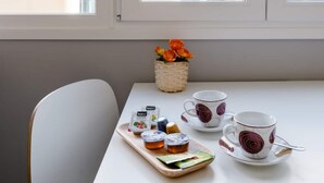Uma mesa com bandeja de comida e duas xícaras com pires, um cesto de flores e uma janela com vista para edifícios.