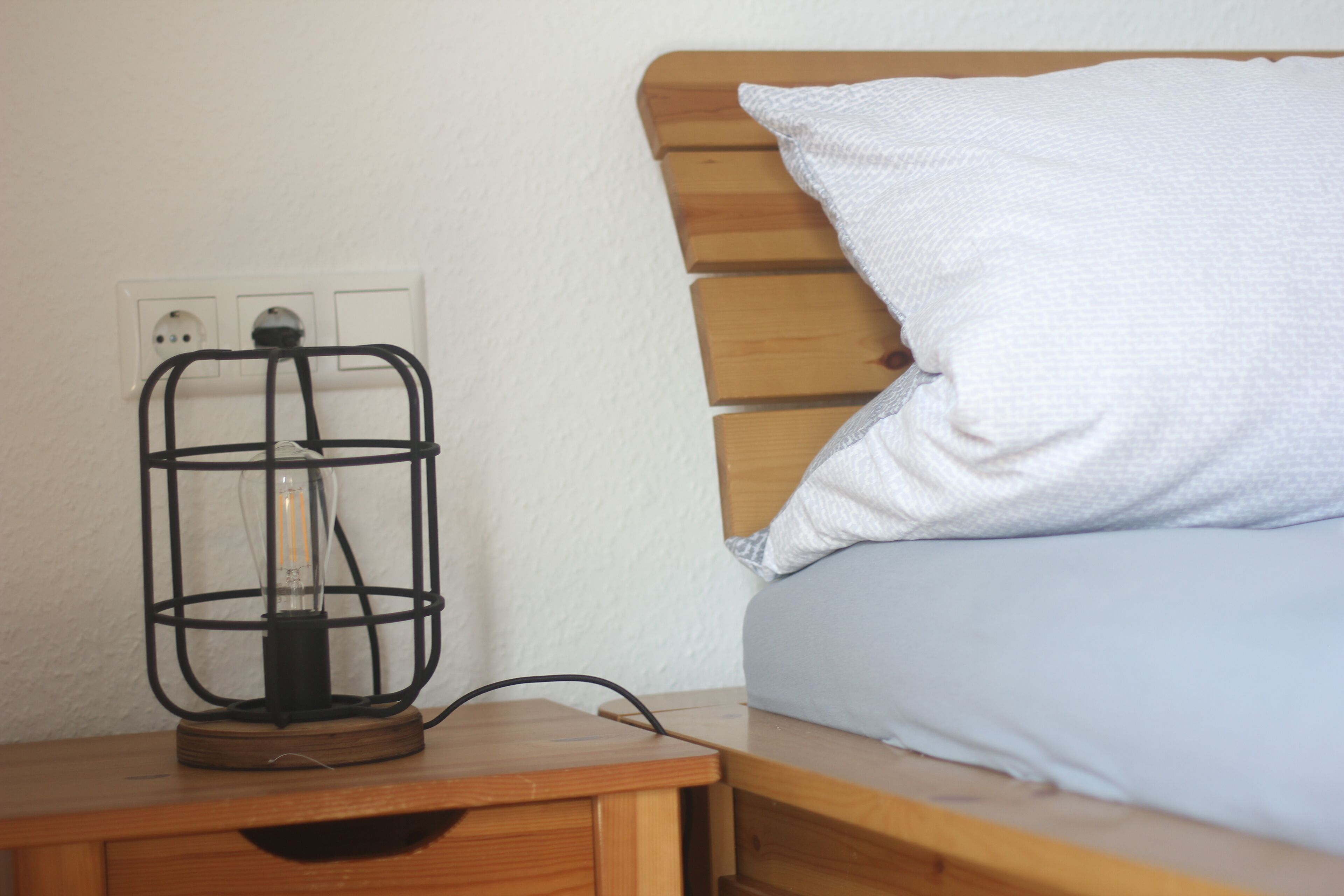 A wooden bedside table with a lamp and a power outlet on the wall.