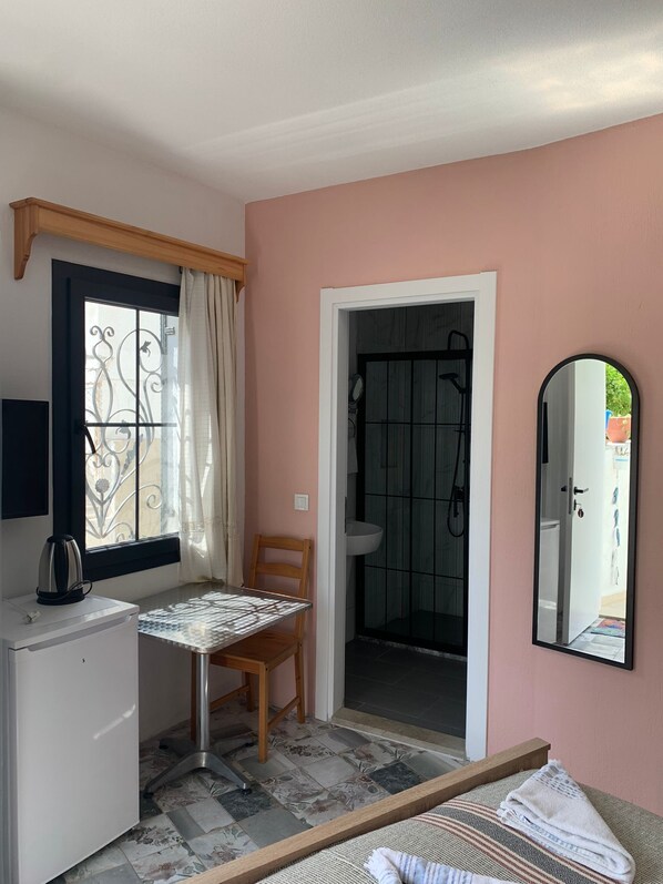 A room with a bed, a small table with a chair, a desk with a kettle, a window with curtains, a bathroom with a shower, and a mirror on the wall.