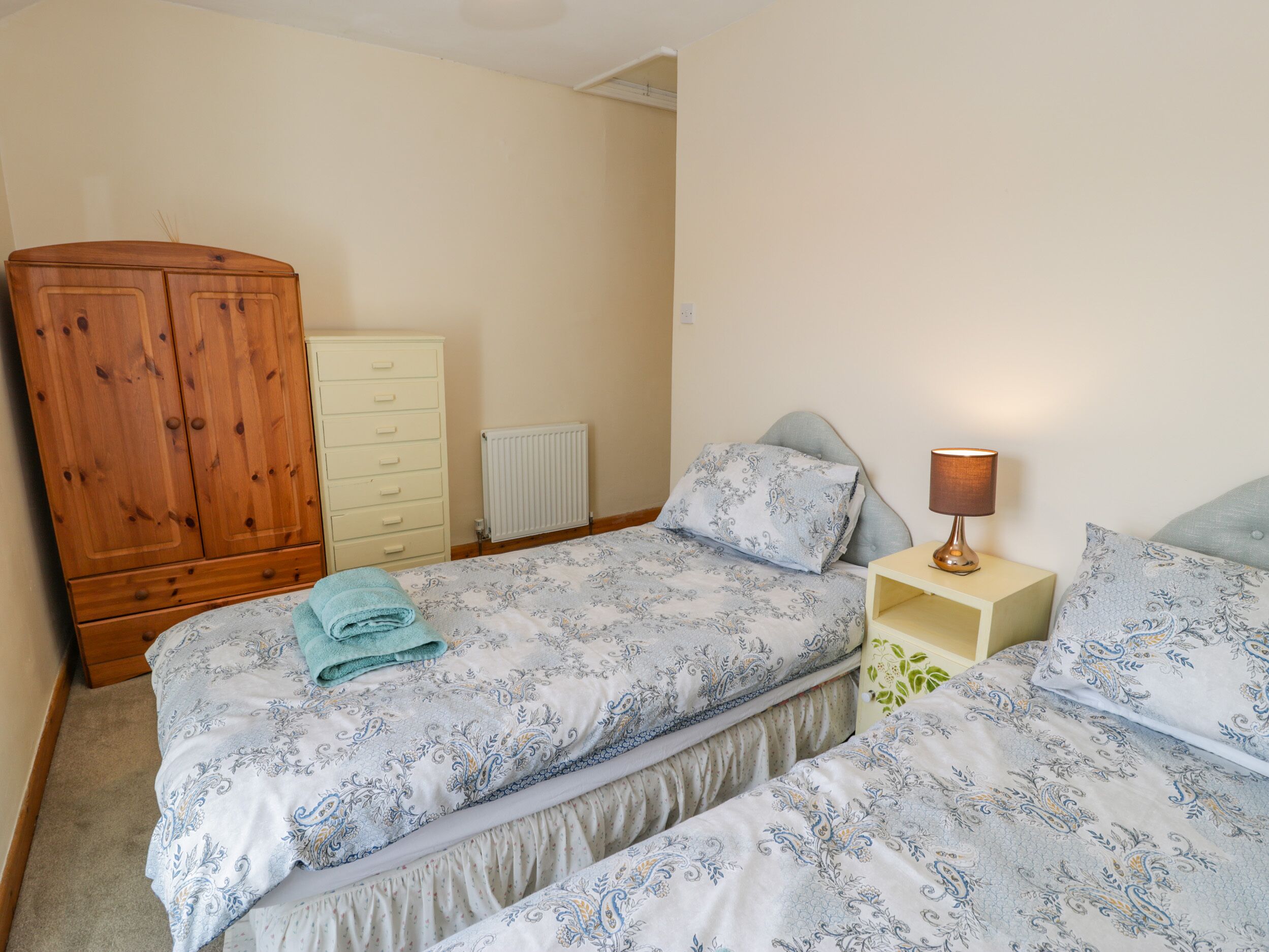 A bedroom with two beds, a wooden wardrobe, a radiator, and a bedside lamp.