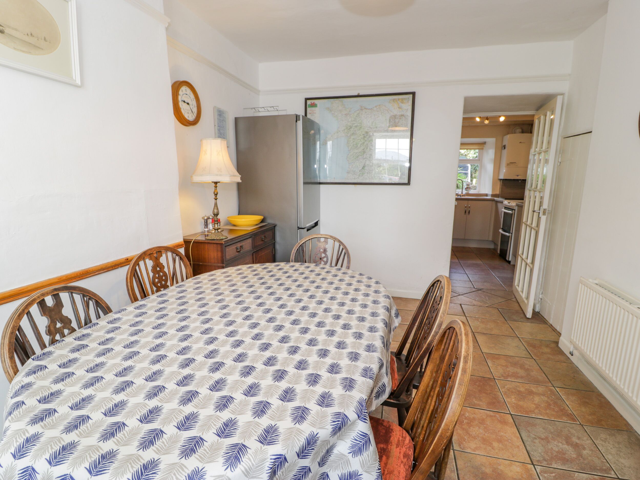 A dining area with a table set for four, a lamp, a map on the wall, and a kitchen in the background.