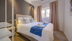 A neatly made bed with a blue blanket, white pillows, and a rolled towel. There is a bedside table with a lamp, a window with curtains, and an air conditioning unit on the wall. - Tritoni harbour suites (Valletta)