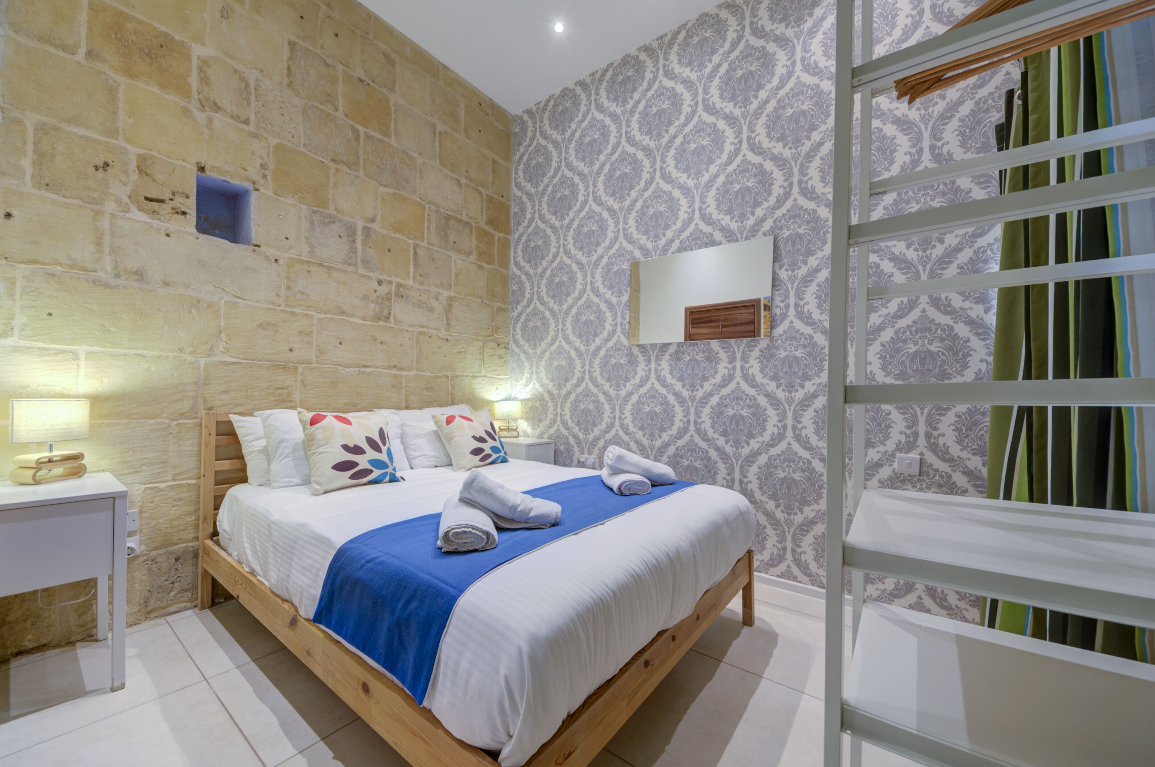 A bedroom with a wooden bed, white bedding, a blue throw, and a nightstand with a lamp.