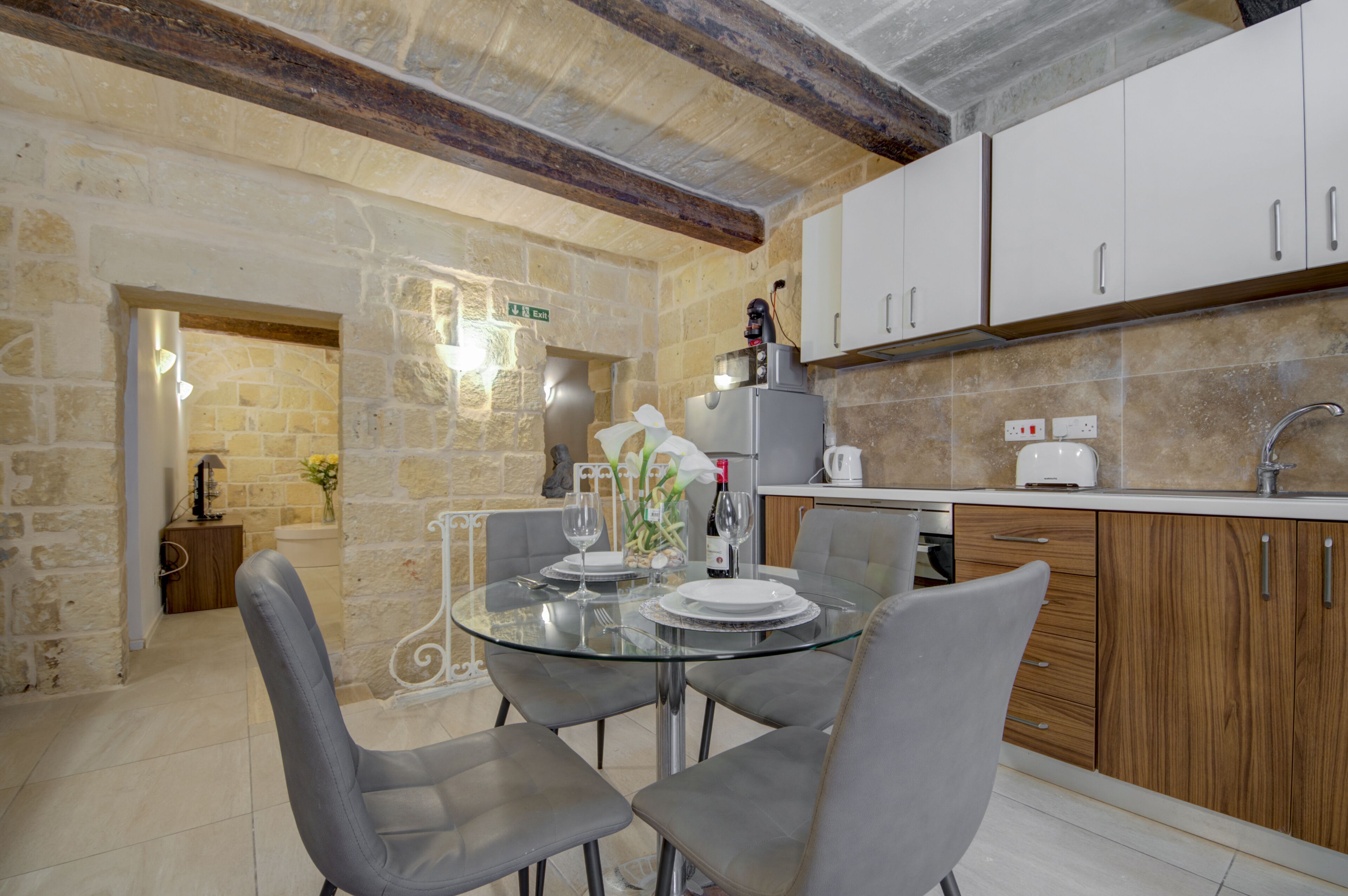 A modern kitchen with a glass dining table, grey chairs, and wooden cabinets.