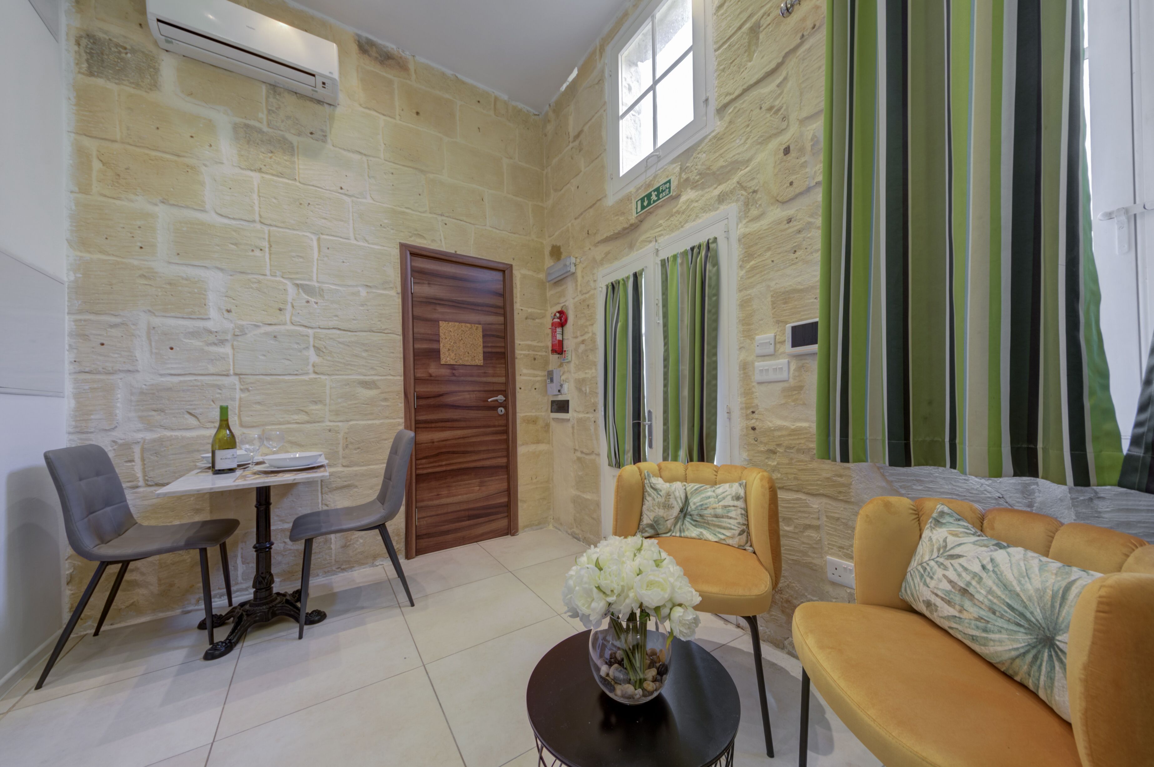 A room with a stone wall, a wooden door, a small table with a bottle and glasses, two chairs, a sofa with cushions, and a window with green curtains.
