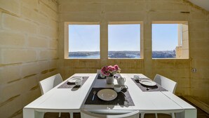 A dining area with a white table set for four, three windows offering a view of the city, and a vase with pink flowers on the table. - Tritoni harbour suites (Valletta)