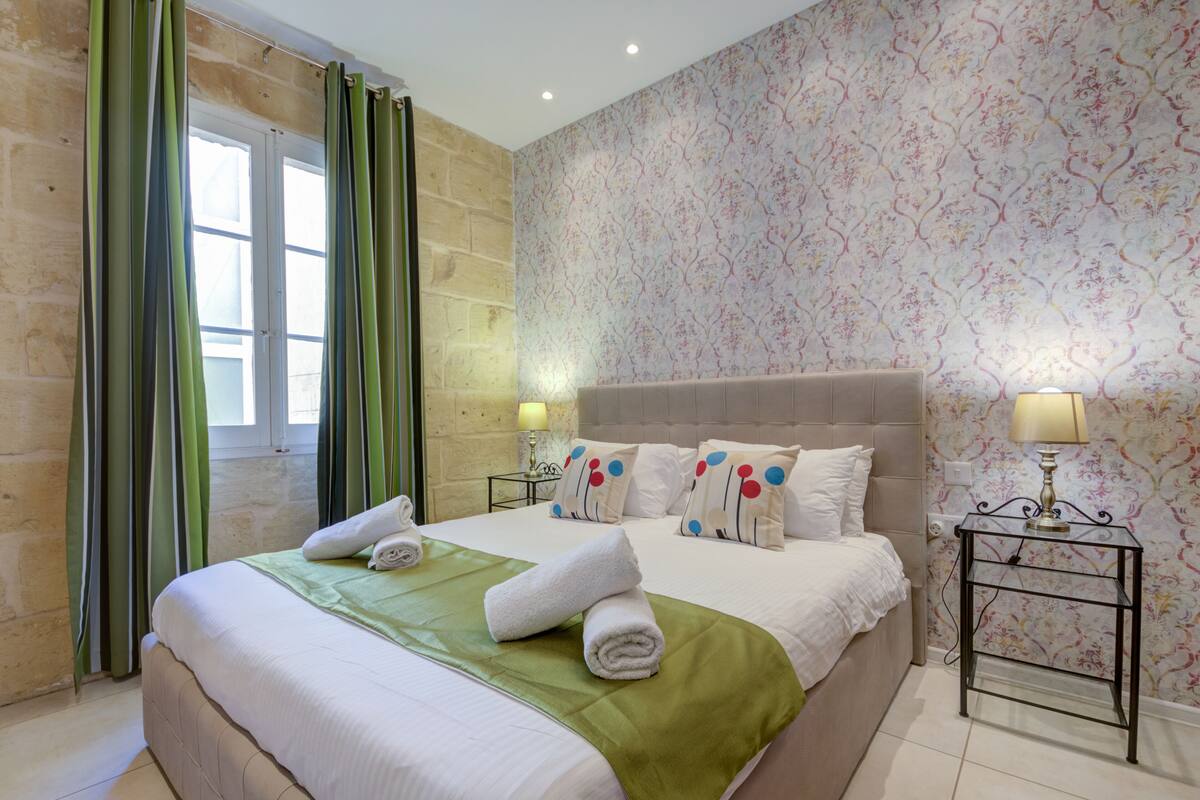 A hotel room with a large bed, green curtains, floral wallpaper, and two bedside lamps.