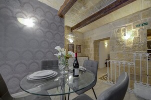 Un comedor con una mesa de vidrio, platos blancos, una botella de vino y un florero con flores. Se observa una pared de piedra y un letrero que indica una salida.