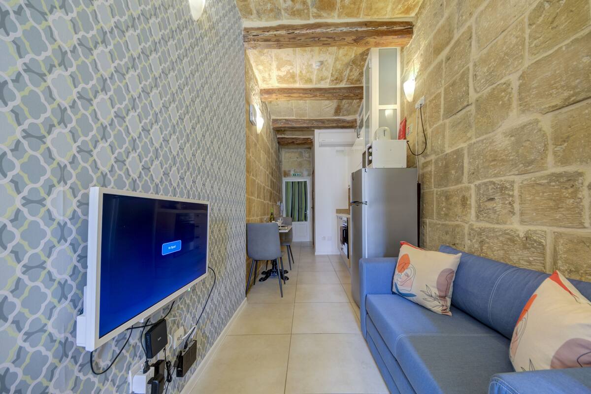 A narrow hallway with a patterned wall, a television, a sofa with decorative pillows, and a kitchen area with a refrigerator.