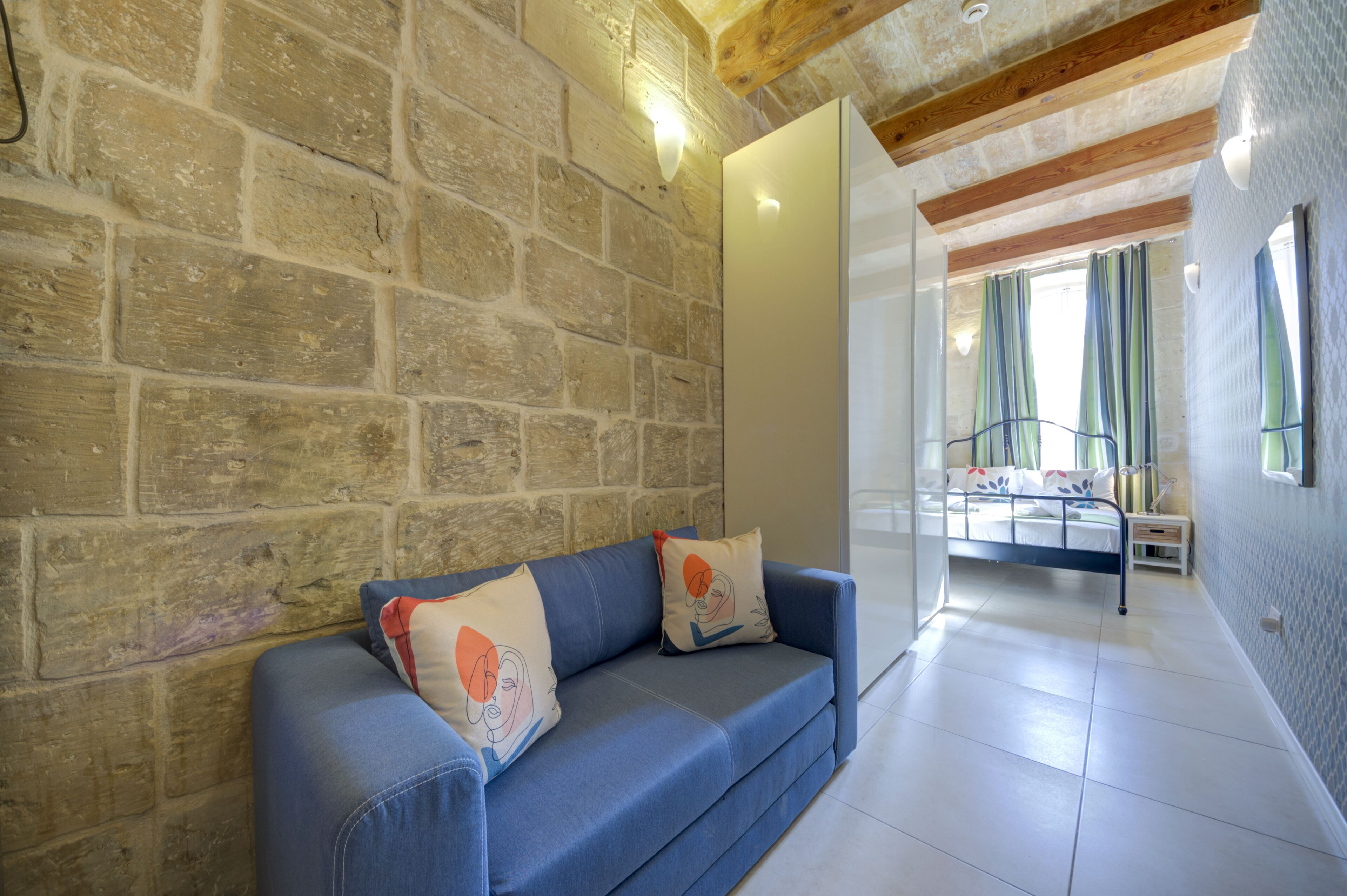 A room with a stone wall, a blue sofa with decorative pillows, a bed with a metal frame, and a window with curtains.