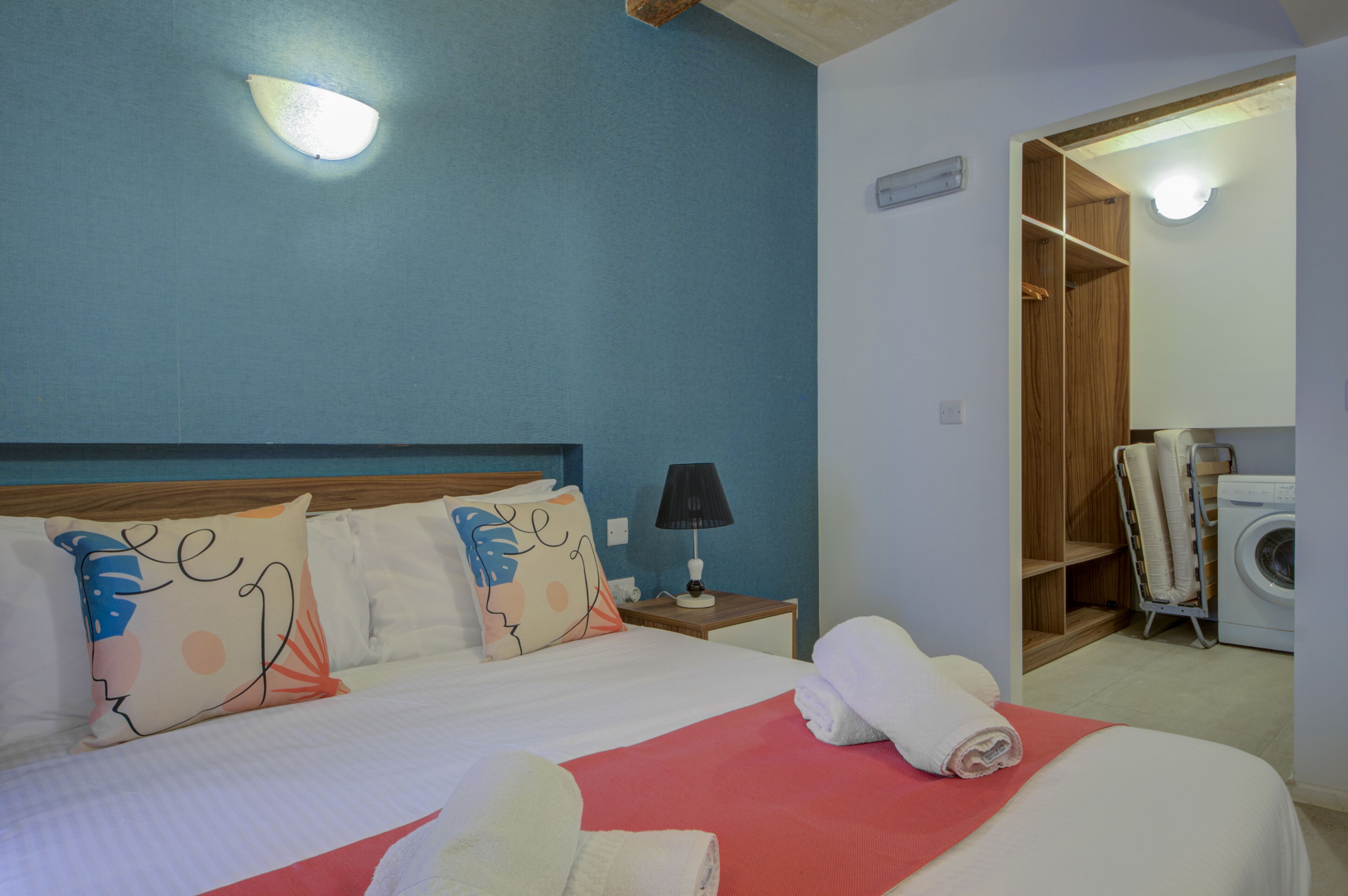 Una habitación de hotel con cama, mesita de noche, lámpara, armario y lavadora.