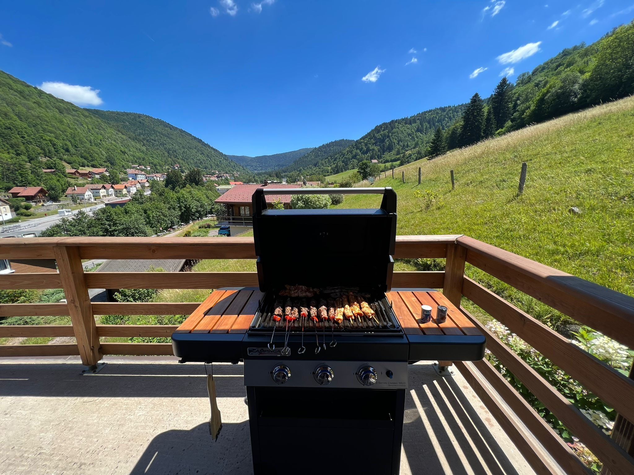 Un barbecue installé sur une terrasse en bois, offrant une vue panoramique sur une vallée et des maisons.