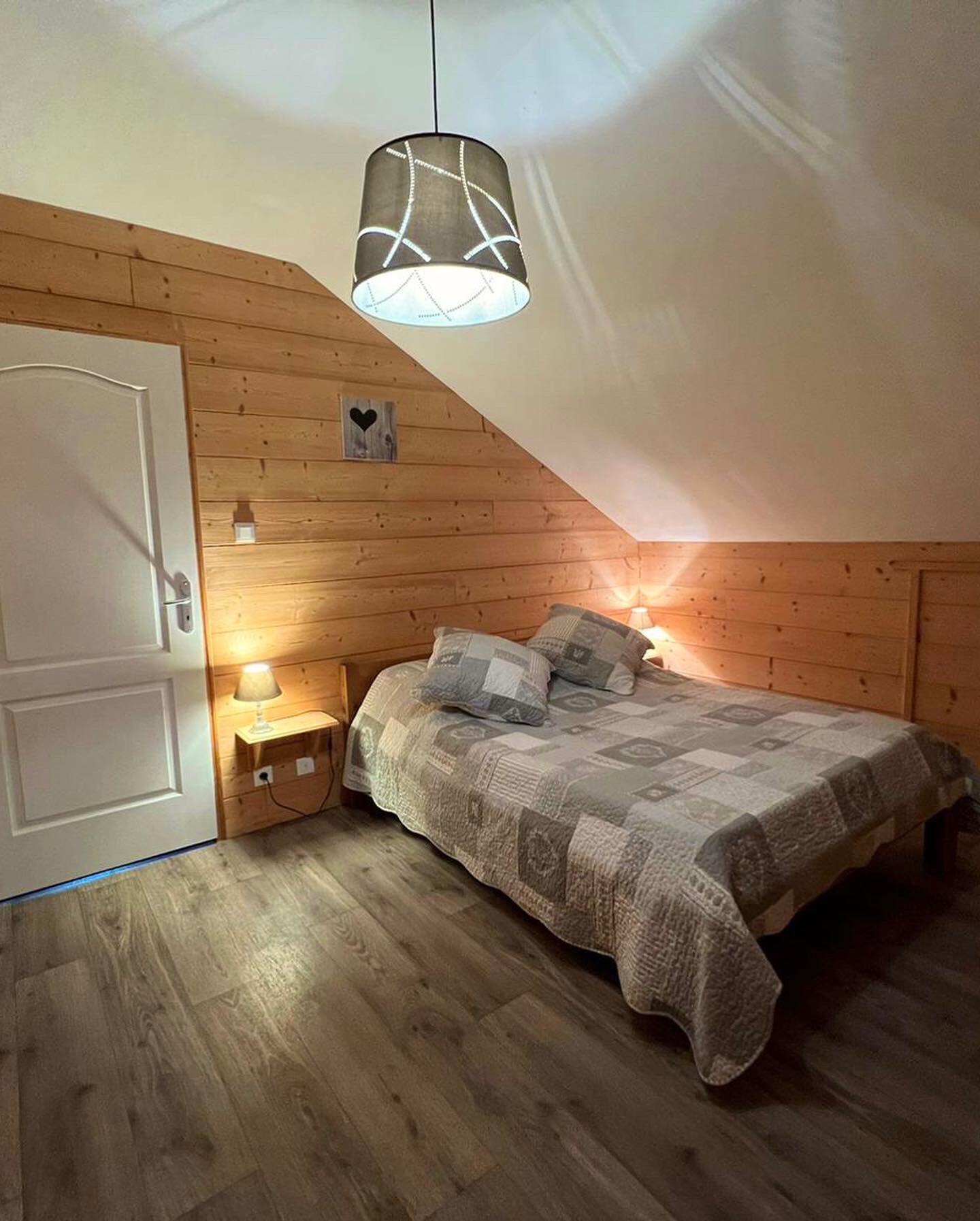 A bedroom with a sloped ceiling, wooden walls, a bed with a patterned blanket, a bedside lamp, and a hanging light fixture.