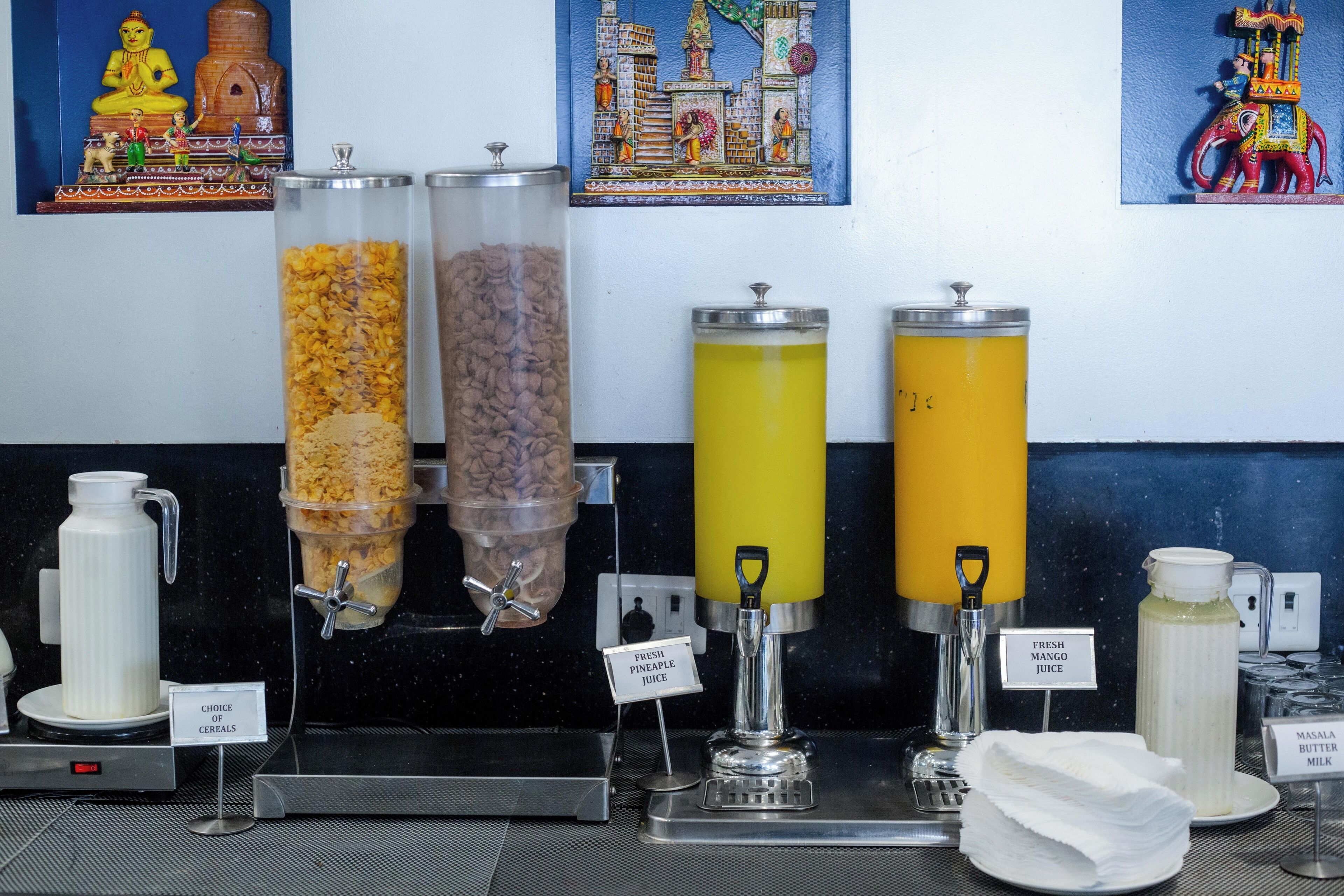 A breakfast bar with various food and drink dispensers, including cereal, coffee, fresh pineapple juice, and mango juice.