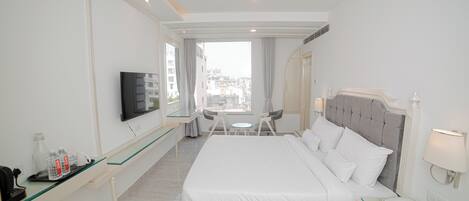 A hotel room with a bed, a TV, a desk with a chair, a small table, and a window with a city view.