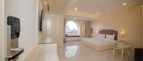 A modern hotel room with a large bed, a bedside table, a seating area, and a view of the city through a large window.