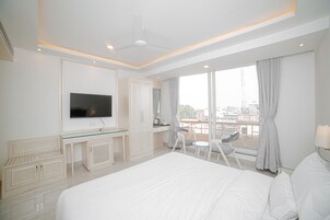 A modern hotel room with a large bed, a TV, a desk with a glass top, and a view of the city through large windows.