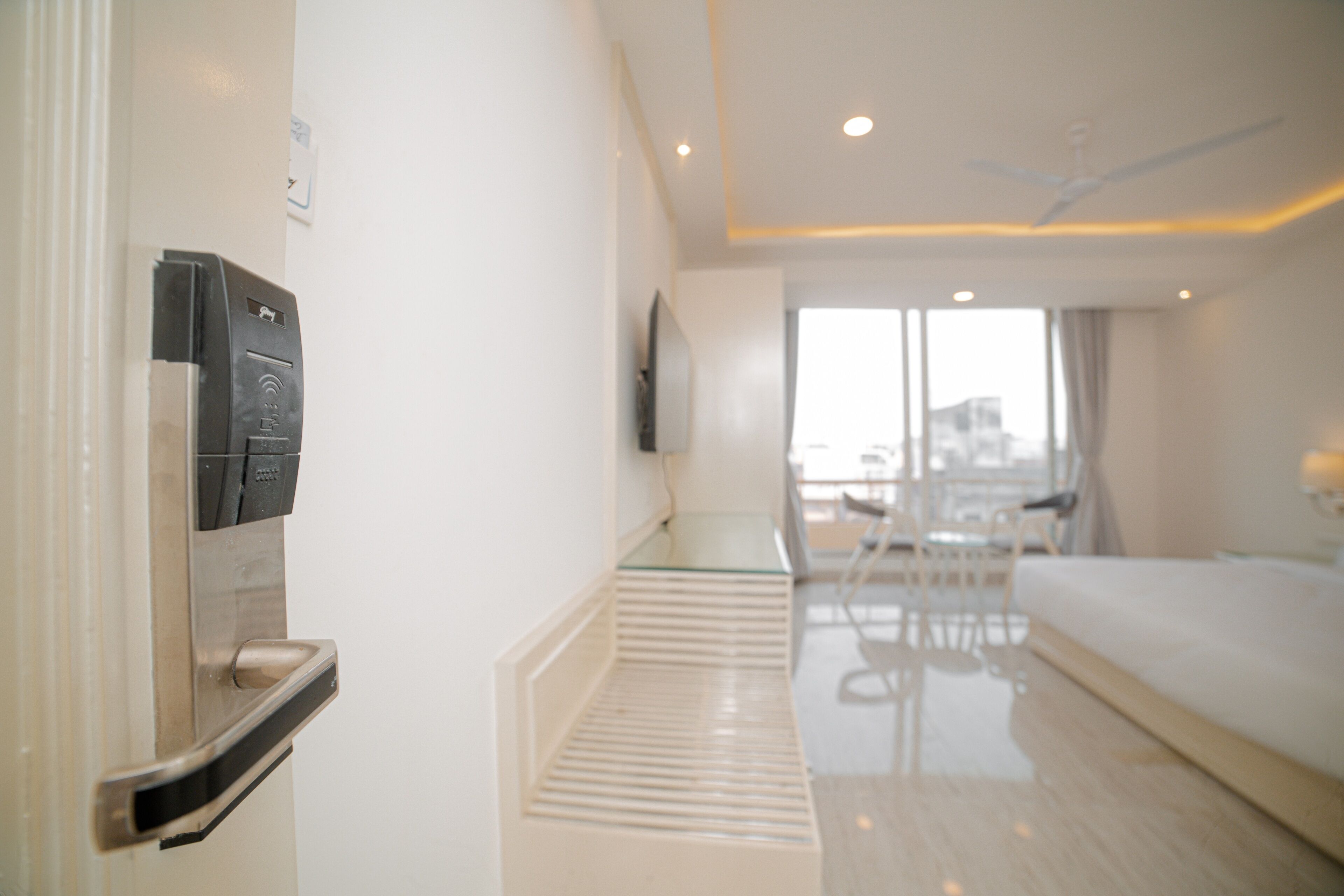 A hotel room with a minibar, a glass table, a bed, and a view of the city through large windows.
