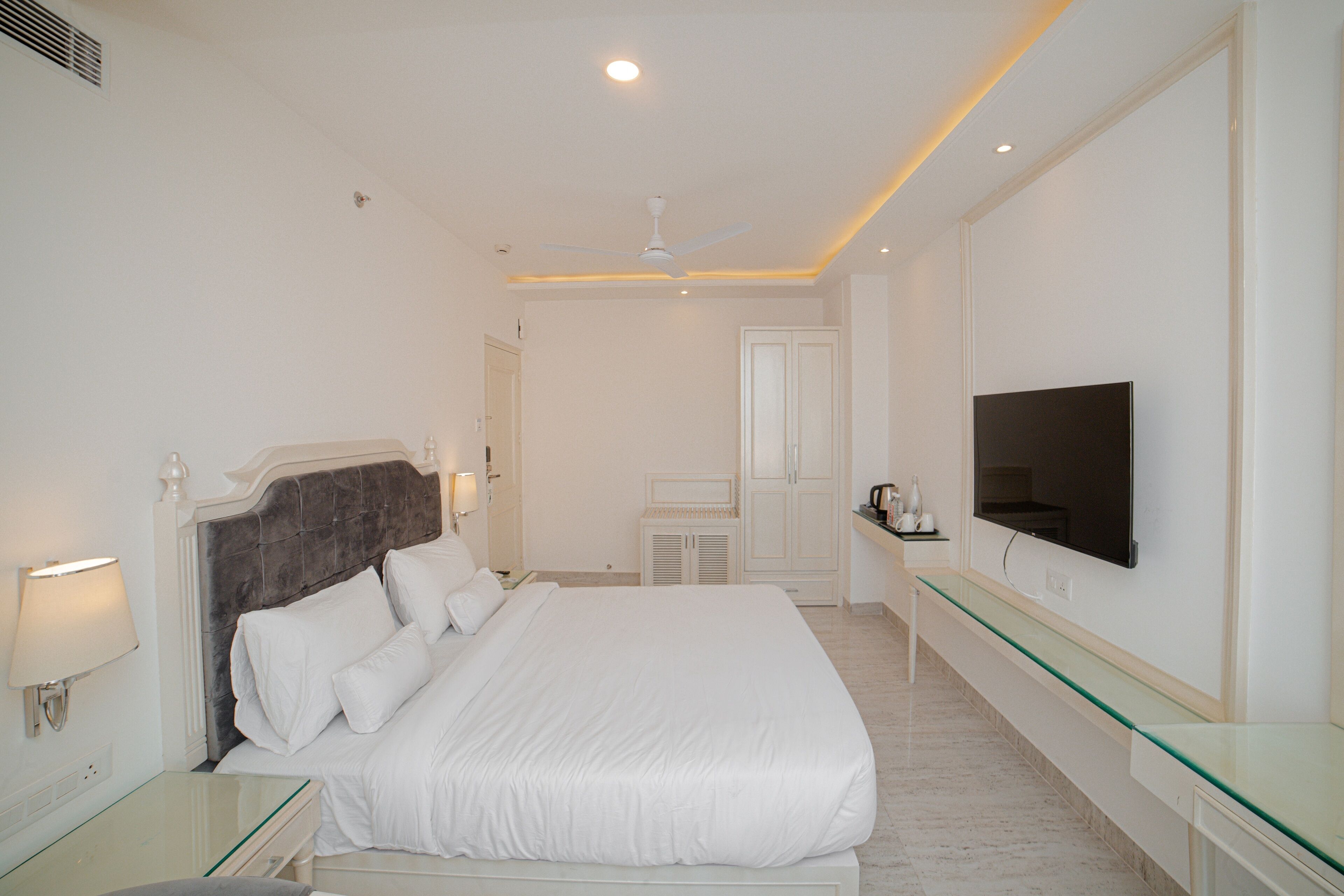 A modern hotel room with a large bed, a flat-screen TV, a glass coffee table, and a ceiling fan.