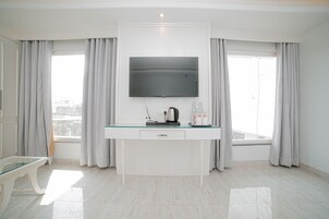 Una habitación de hotel moderna con un televisor de pantalla plana grande montado en la pared, una mesa de vidrio con una tetera y tazas de café, y vistas a la ciudad a través de amplios ventanales con cortinas grises.