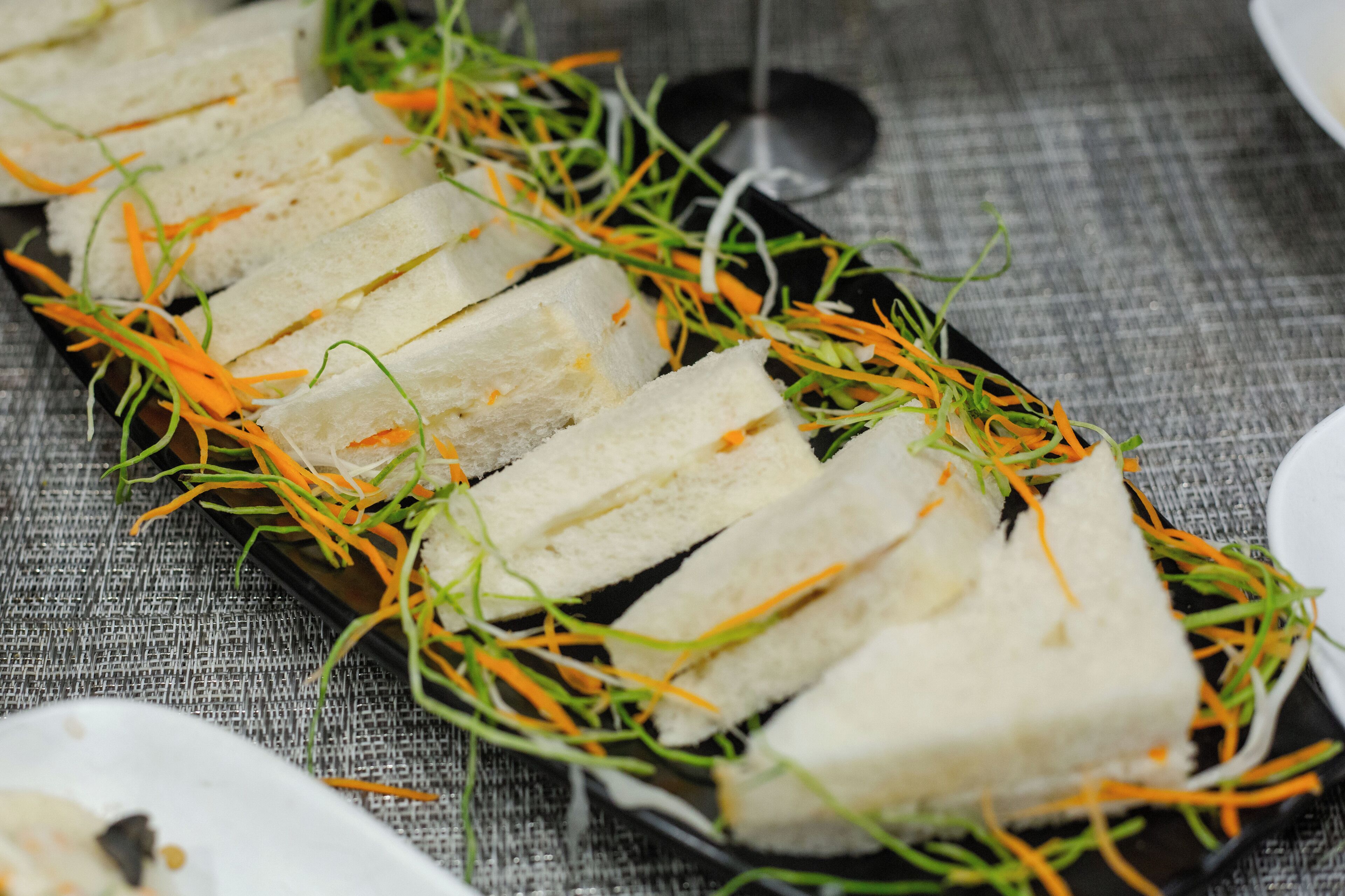 A tray of sandwiches garnished with shredded carrots and greens, placed on a textured tablecloth.