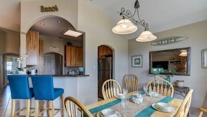 Un comedor con una mesa larga, taburetes de bar azules, sillas de madera y una cocina al fondo.
