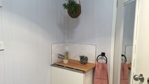 Et badeværelse med en hvid flisevæg, en træhylde med en plante og en beholder, en håndklædering med et pink håndklæde, og en dør med et rundt håndtag.