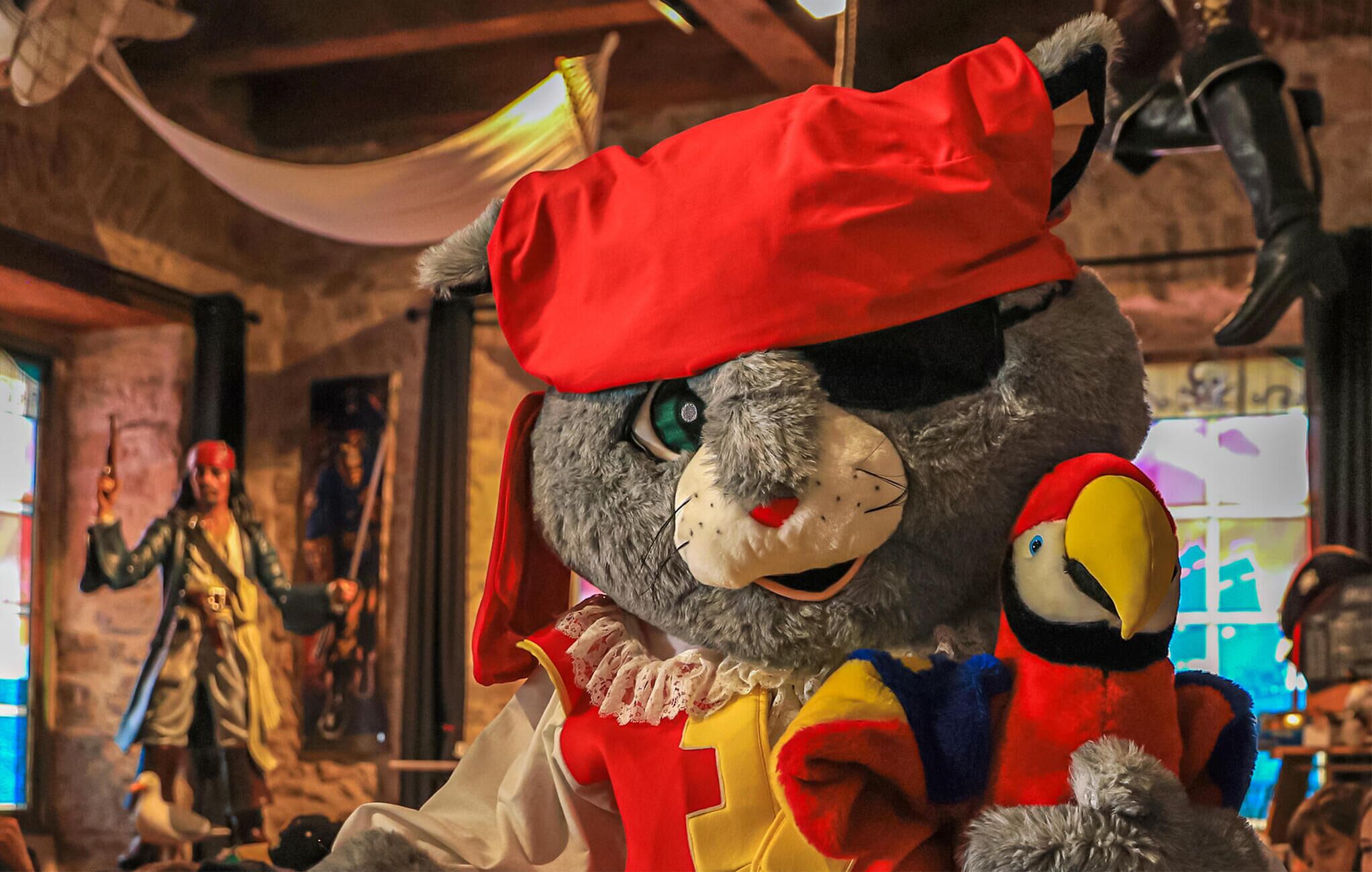 Un personaje de peluche con sombrero y vestimenta roja, sosteniendo un juguete más pequeño, en un ambiente rústico y con una figura al fondo.