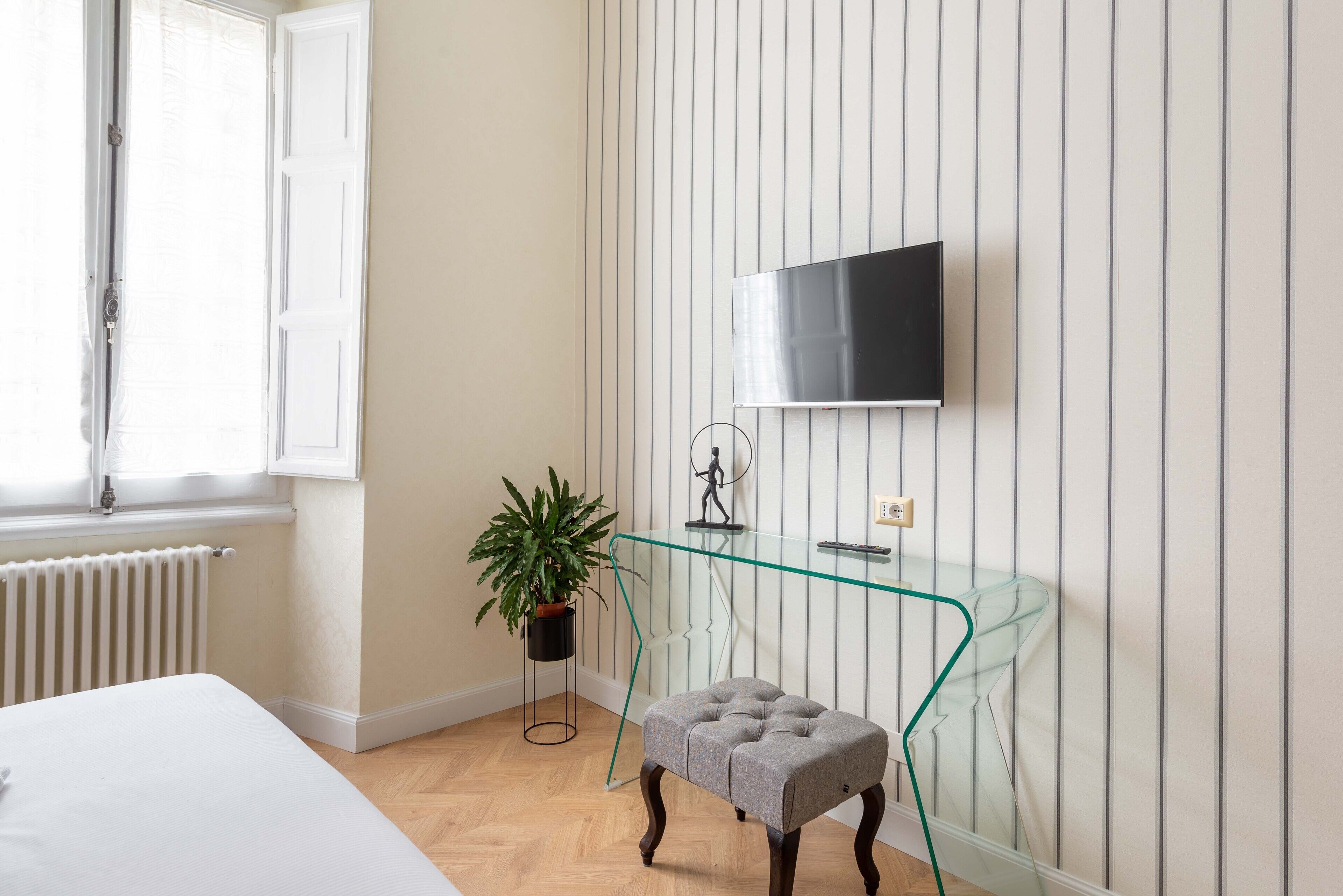 Un dormitorio con cama, un escritorio de vidrio, un reposapiés gris, una planta en maceta y un televisor montado.