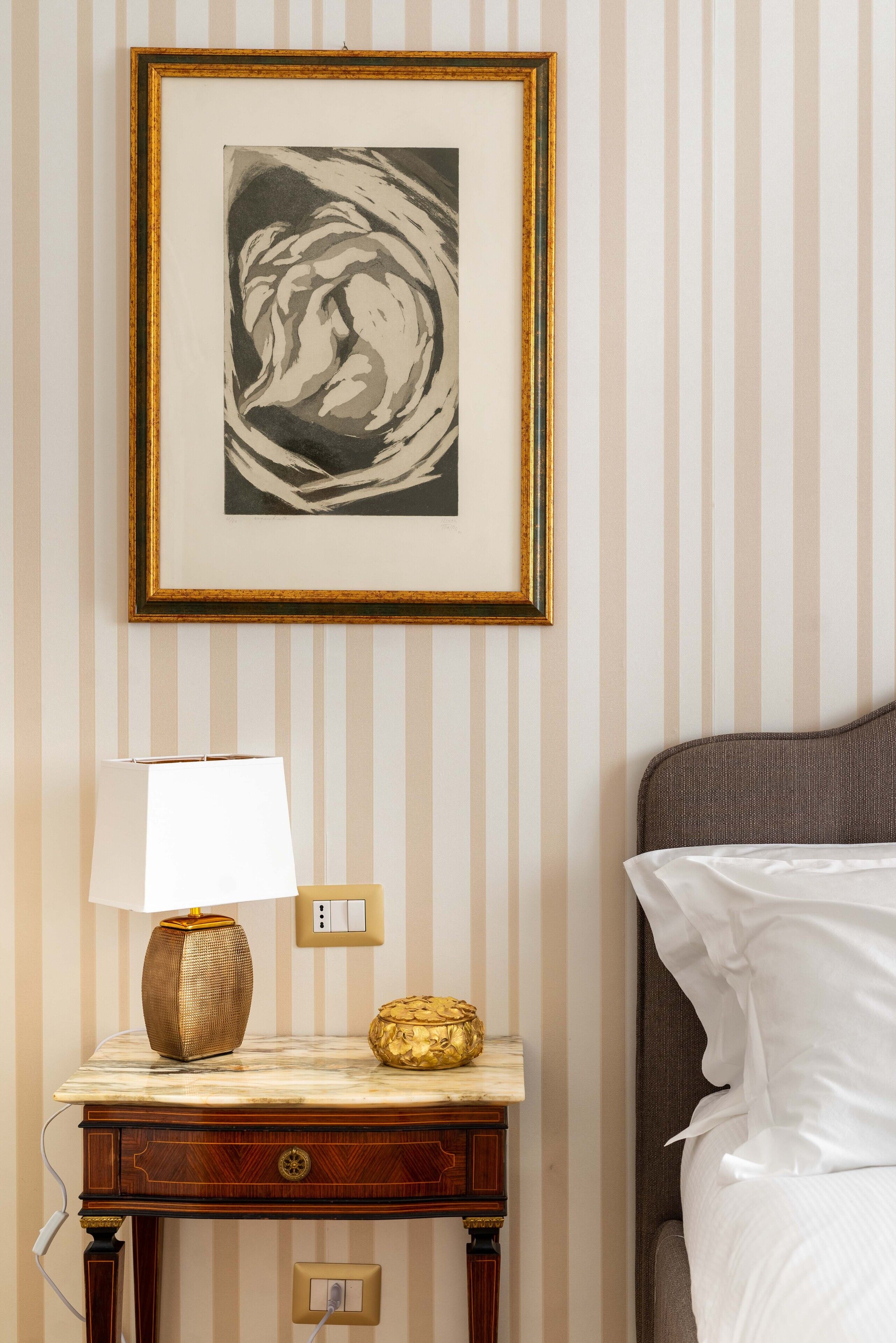 A neatly arranged hotel room with a framed artwork on the wall, a bedside table with a lamp and decorative items, and a bed with white linens.
