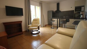 A living room with a sofa, a chair, a TV mounted on the wall, and a kitchen area with a microwave and oven. - Villa Capucine (Flayosc)