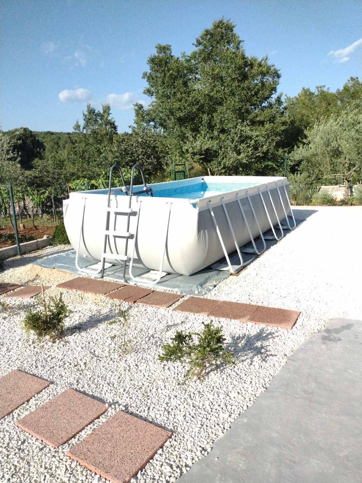 Una piscina con escalera, rodeada de grava y plantas pequeñas, ubicada en una zona ajardinada con árboles al fondo.