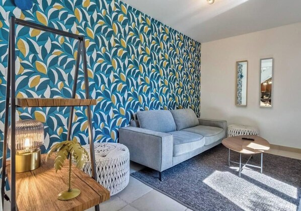A living room with a patterned wallpaper, a grey sofa, a round coffee table, and a small side table with a lamp. - La Cachette du Lac – Near the Lake (MENTHON-SAINT-BERNARD)