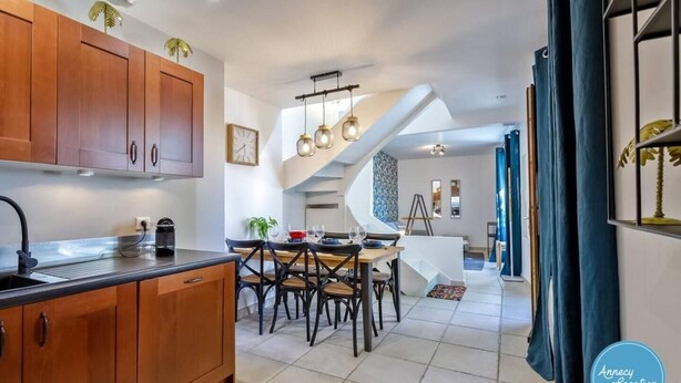 A modern kitchen with wooden cabinets, a dining area with a white table and chairs, and a staircase leading to another level. - La Cachette du Lac – Near the Lake (MENTHON-SAINT-BERNARD)