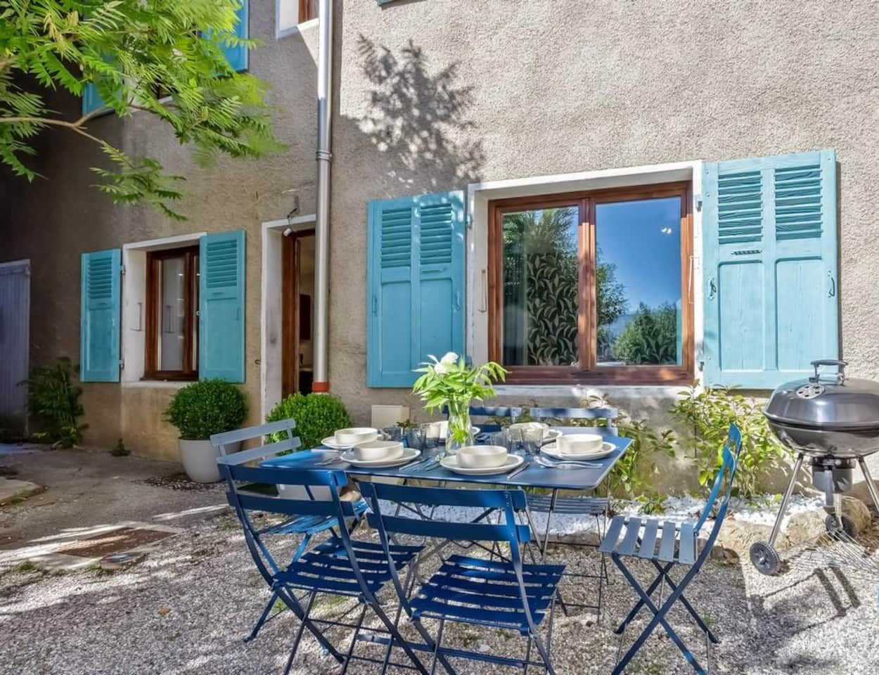 A patio with a table set for four, blue shutters, a grill, and a view of the surrounding area.