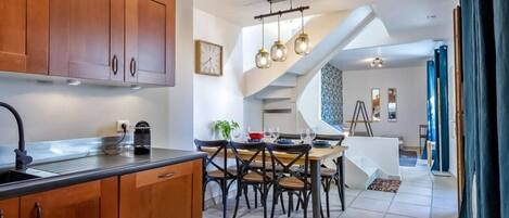 Una cocina moderna con armarios de madera, un comedor con una mesa y sillas blancas, y una escalera que sube a otro nivel.