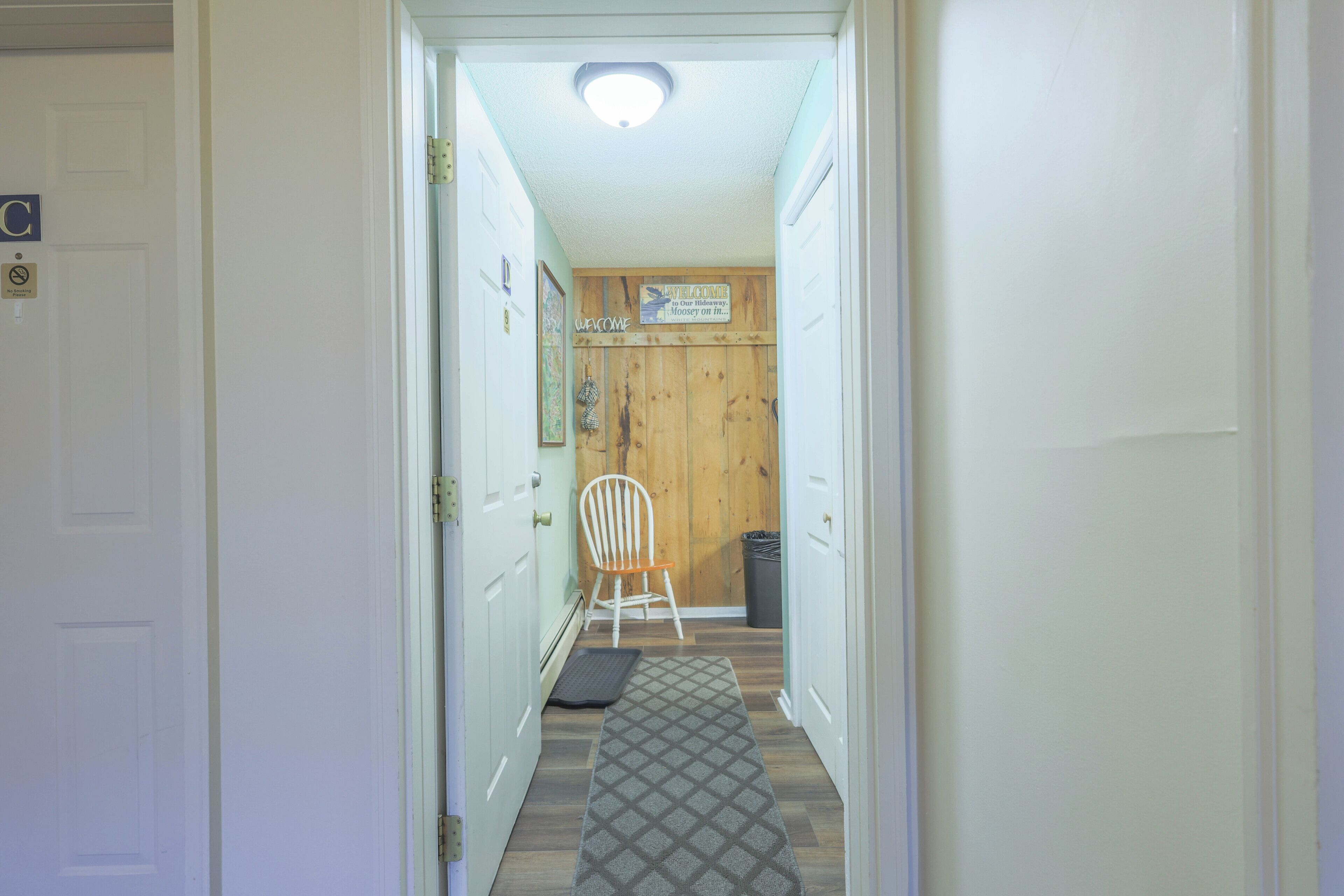A hallway with a wooden door, a welcome sign, a chair, a trash bin, and a mat.