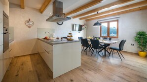Cocina moderna con una isla central, vigas de madera, zona de comedor con sillas, televisor y ventana con vistas.