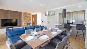 Una sala moderna con un televisor grande, sofás azules, una mesa de comedor preparada para cuatro personas y una cocina con estufa y armarios.