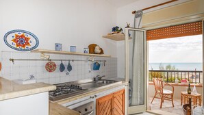 Une cuisine équipée d’une cuisinière à gaz, d’un évier et de meubles en bois. Un balcon avec des chaises en osier offre une vue sur la mer.