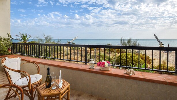 Un balcon avec des fauteuils en osier, une petite table avec une bouteille et des verres, un vase de fleurs et une vue sur la plage et l’océan.