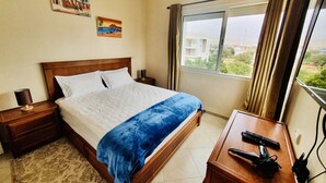 Ein Hotelzimmer mit einem Bett, einem Nachttisch, einem Schreibtisch mit Tastatur, einer Lampe und einem Fenster mit Vorhängen.