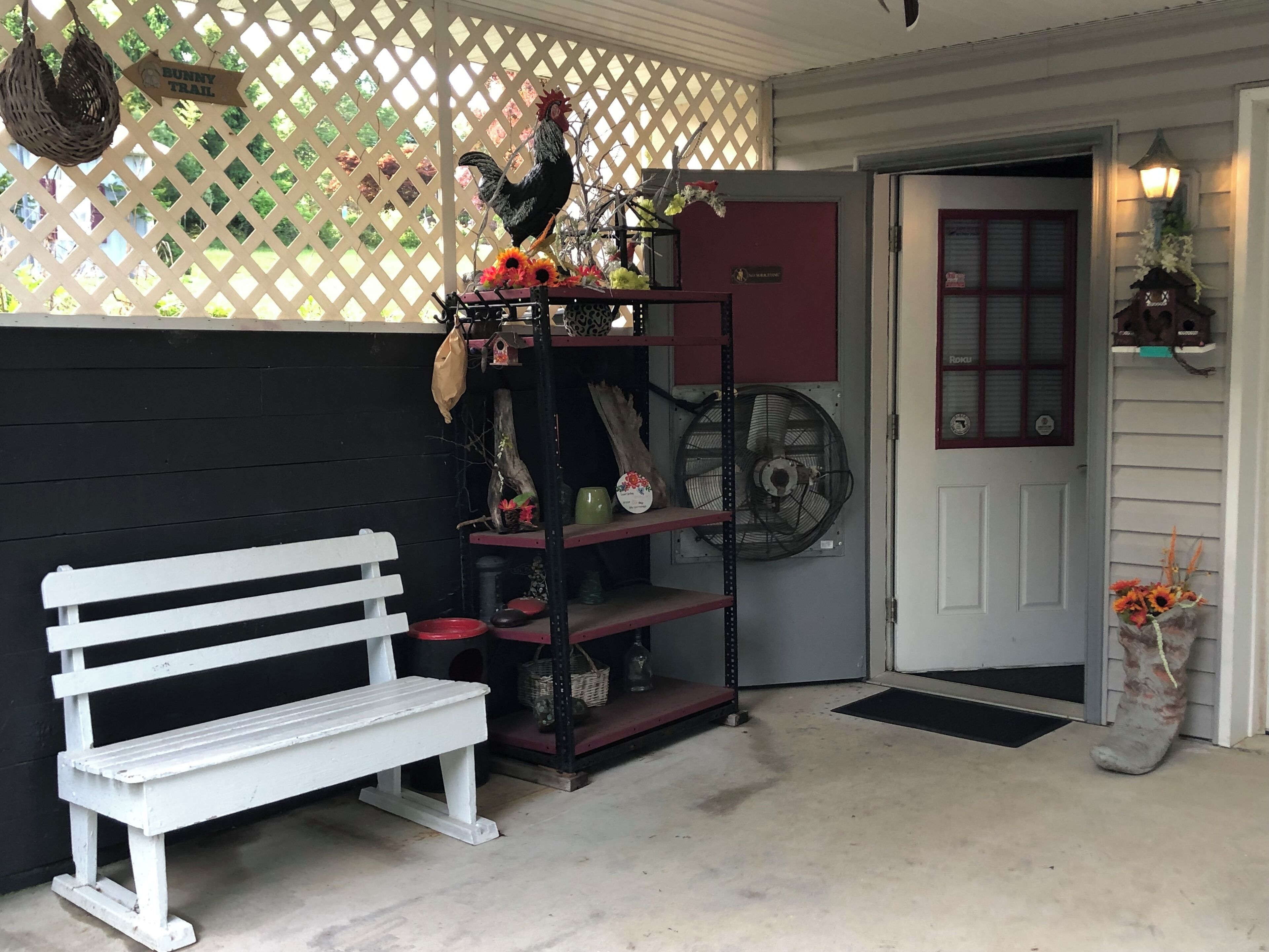 A white bench, a rooster decoration, a fan, and a door with a doormat.