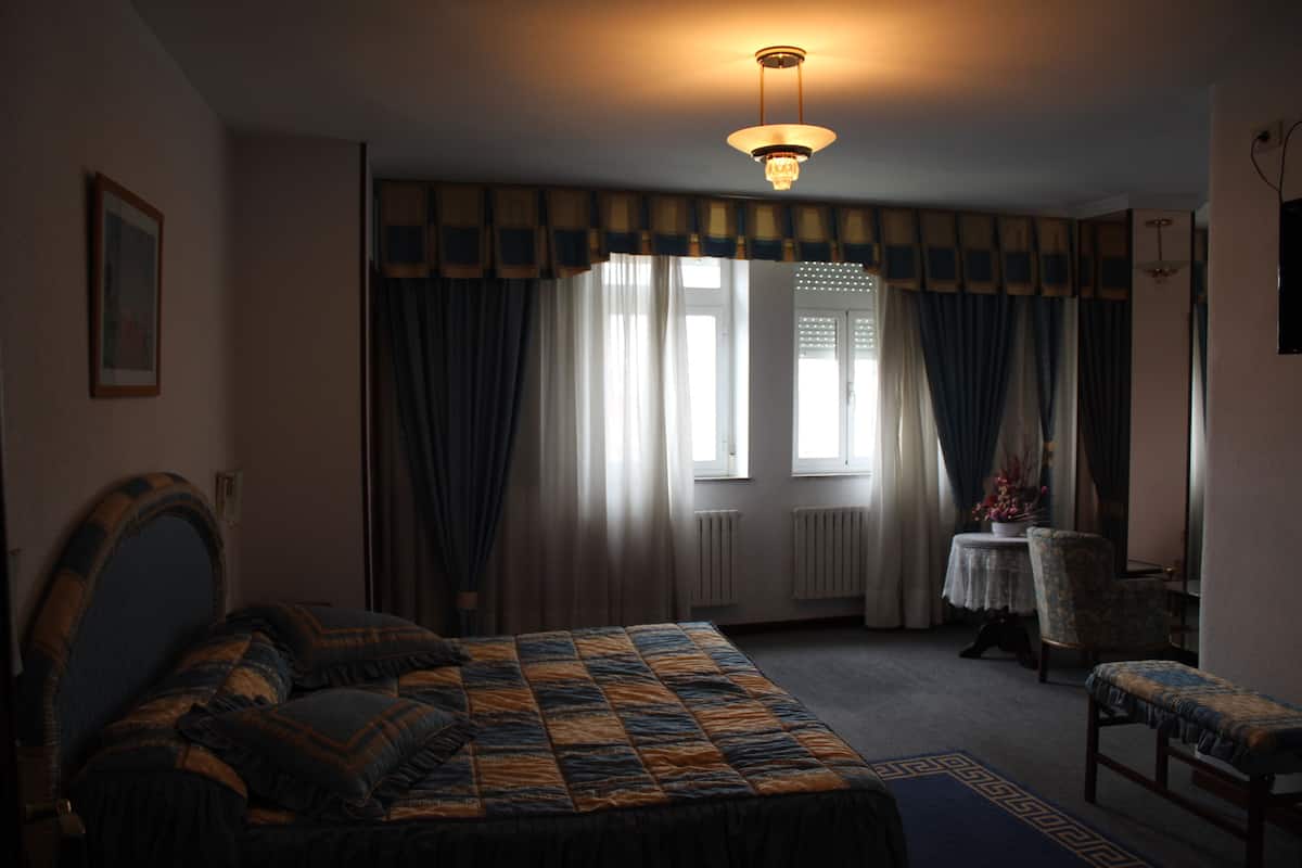A bedroom with a bed, a chair, a table with a vase, and a window with curtains.
